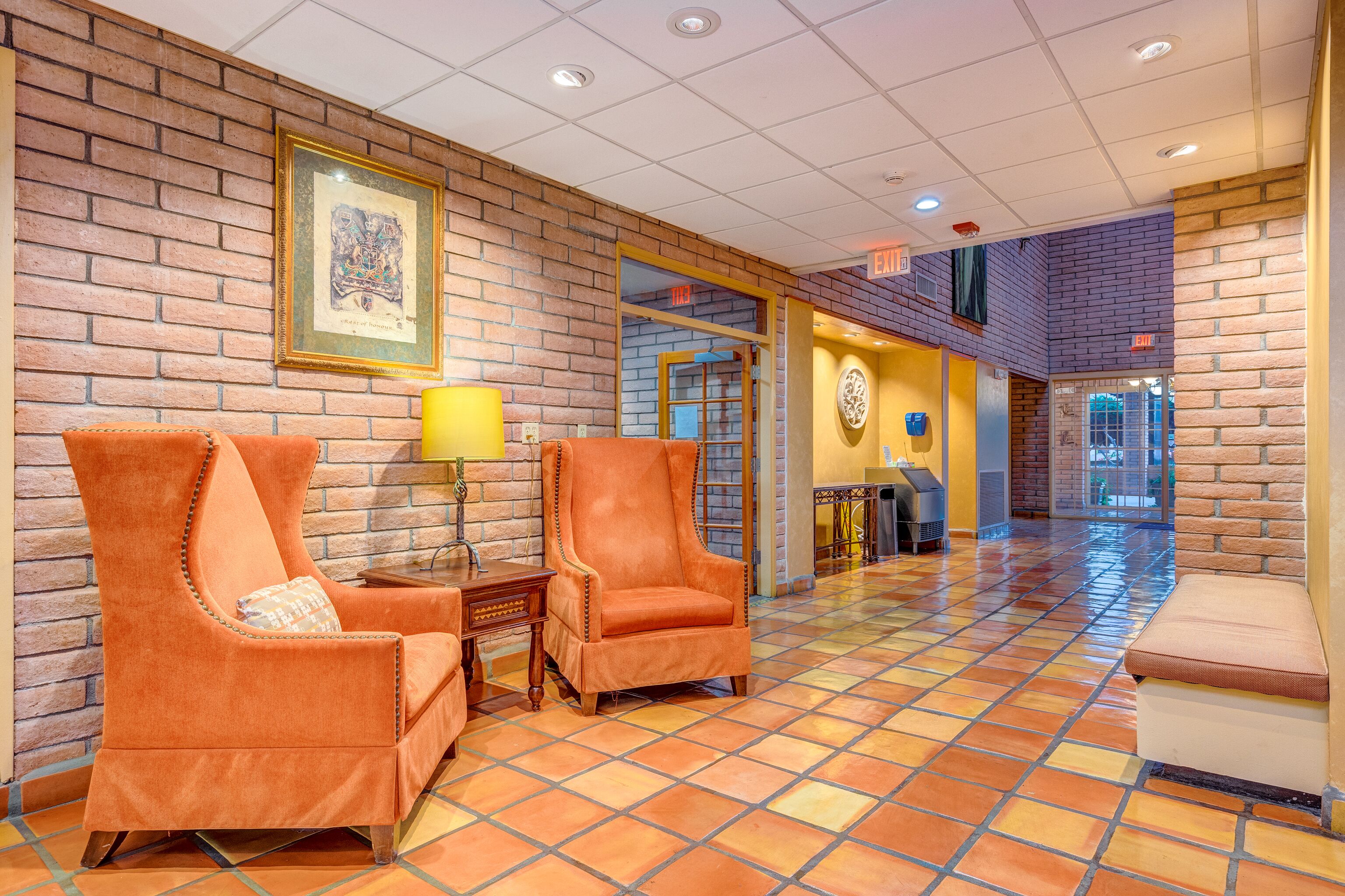 A lobby with brick walls, two orange armchairs, a small table with a lamp, and a bench.