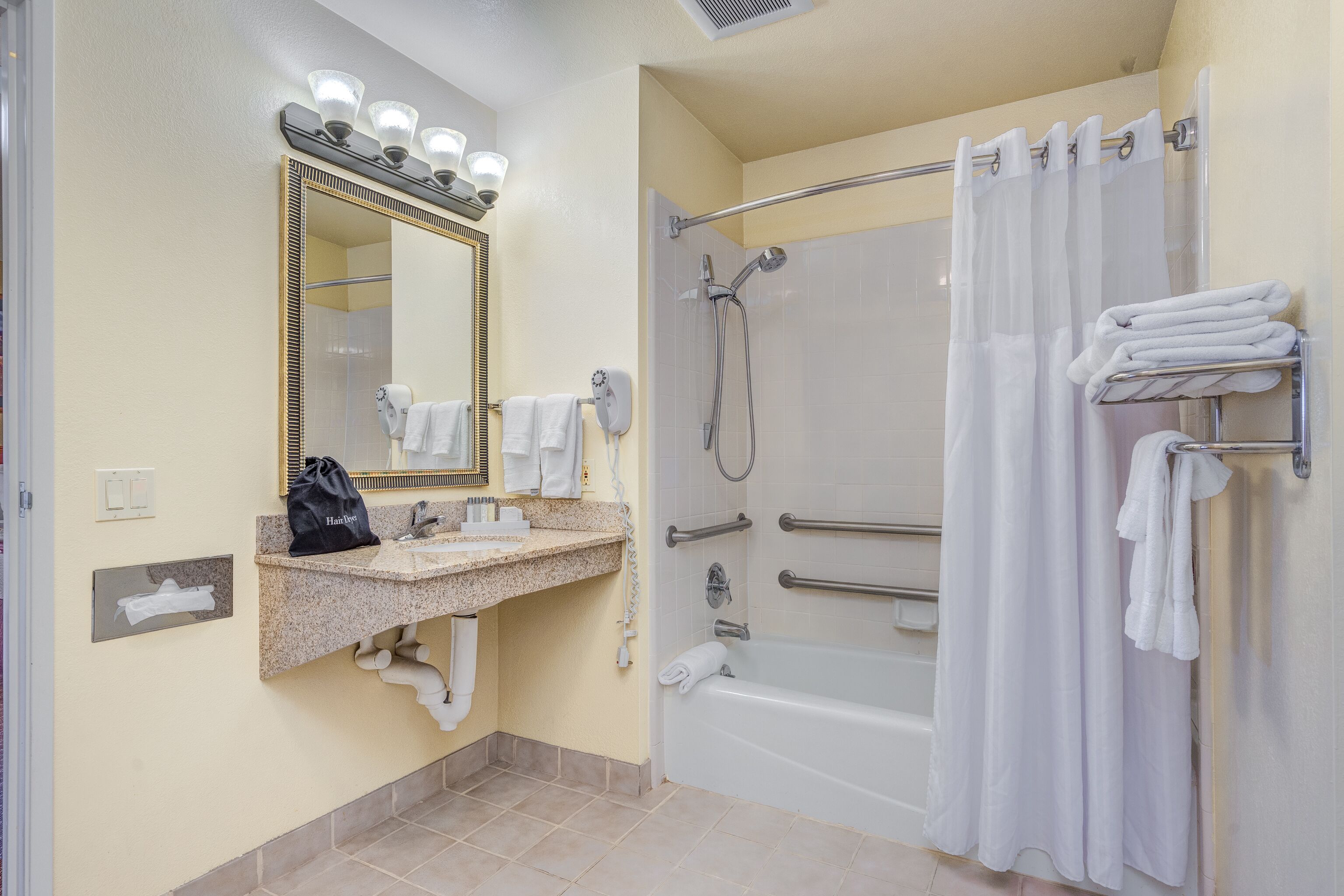 Een badkamer met een wastafel, spiegel, handdoekrek, douche en een zeepdispenser.
