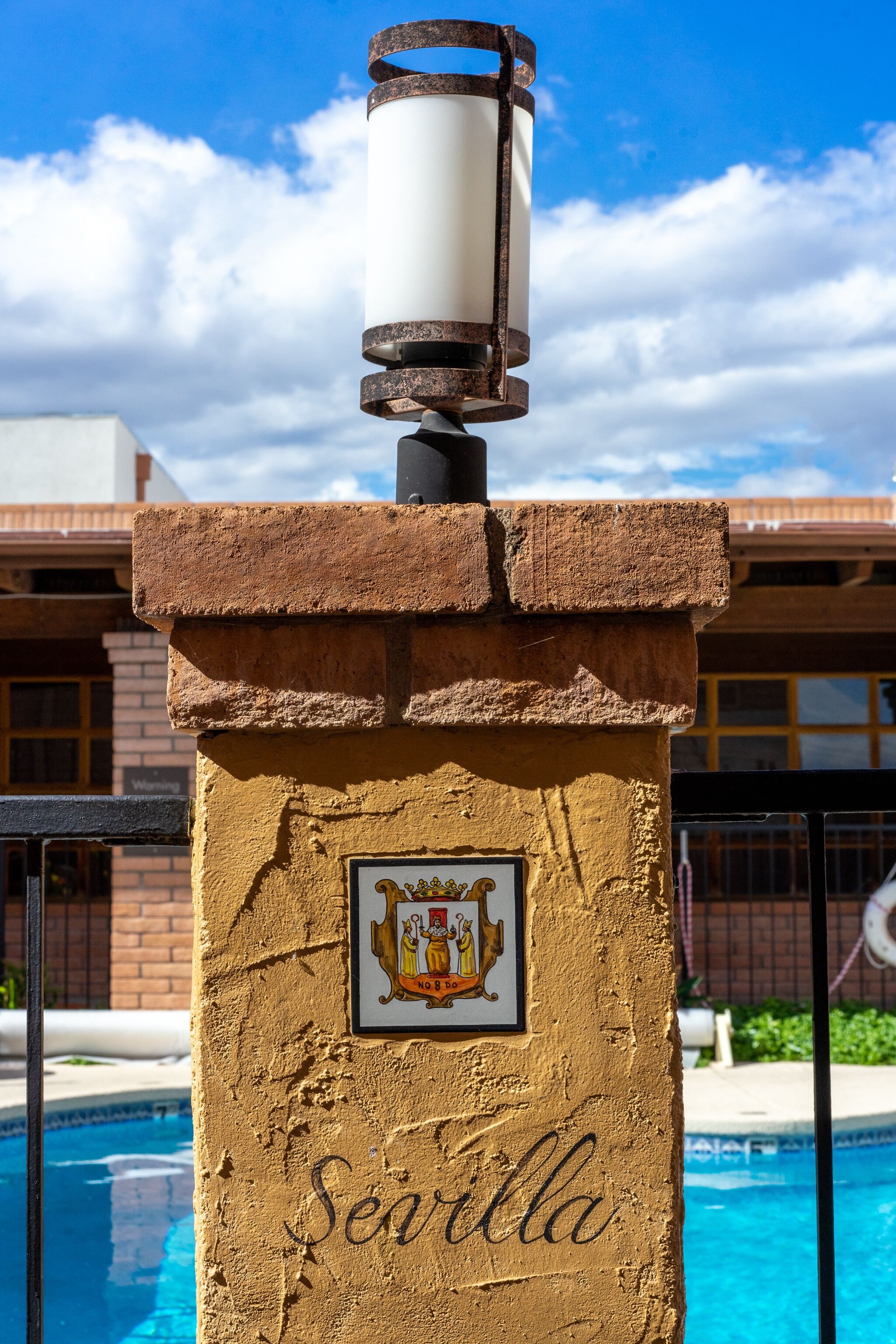 Un pilar de piedra con la palabra "Sevilla" y un escudo, junto a una piscina.