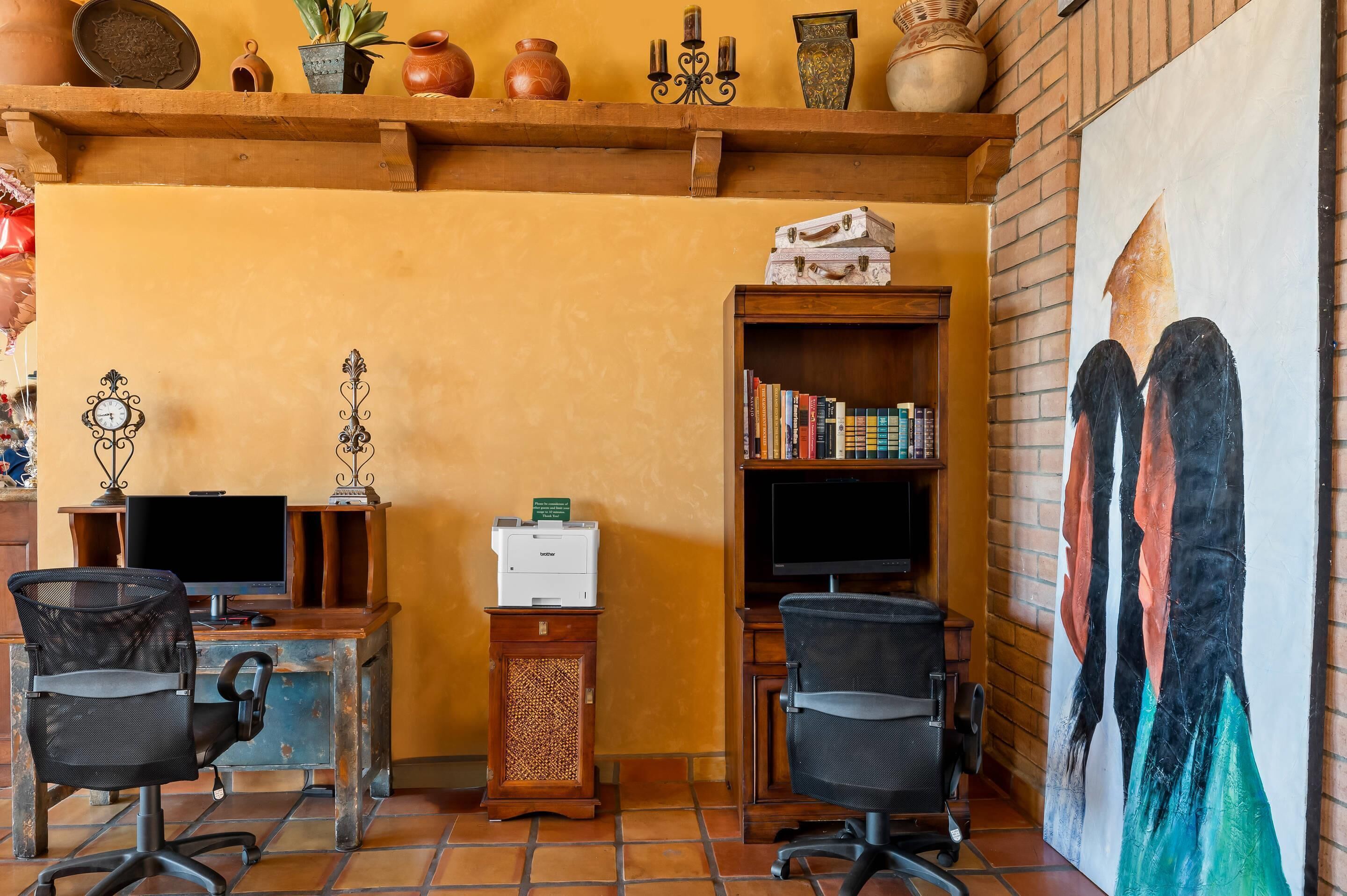 A room with a wooden desk, two chairs, a bookshelf, a printer, and a painting on the wall.