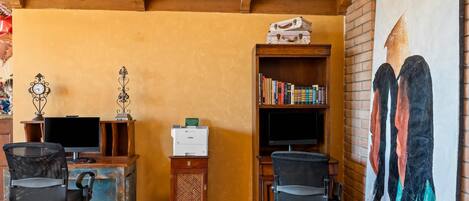 A room with a wooden desk, two chairs, a bookshelf, a printer, and a painting on the wall.