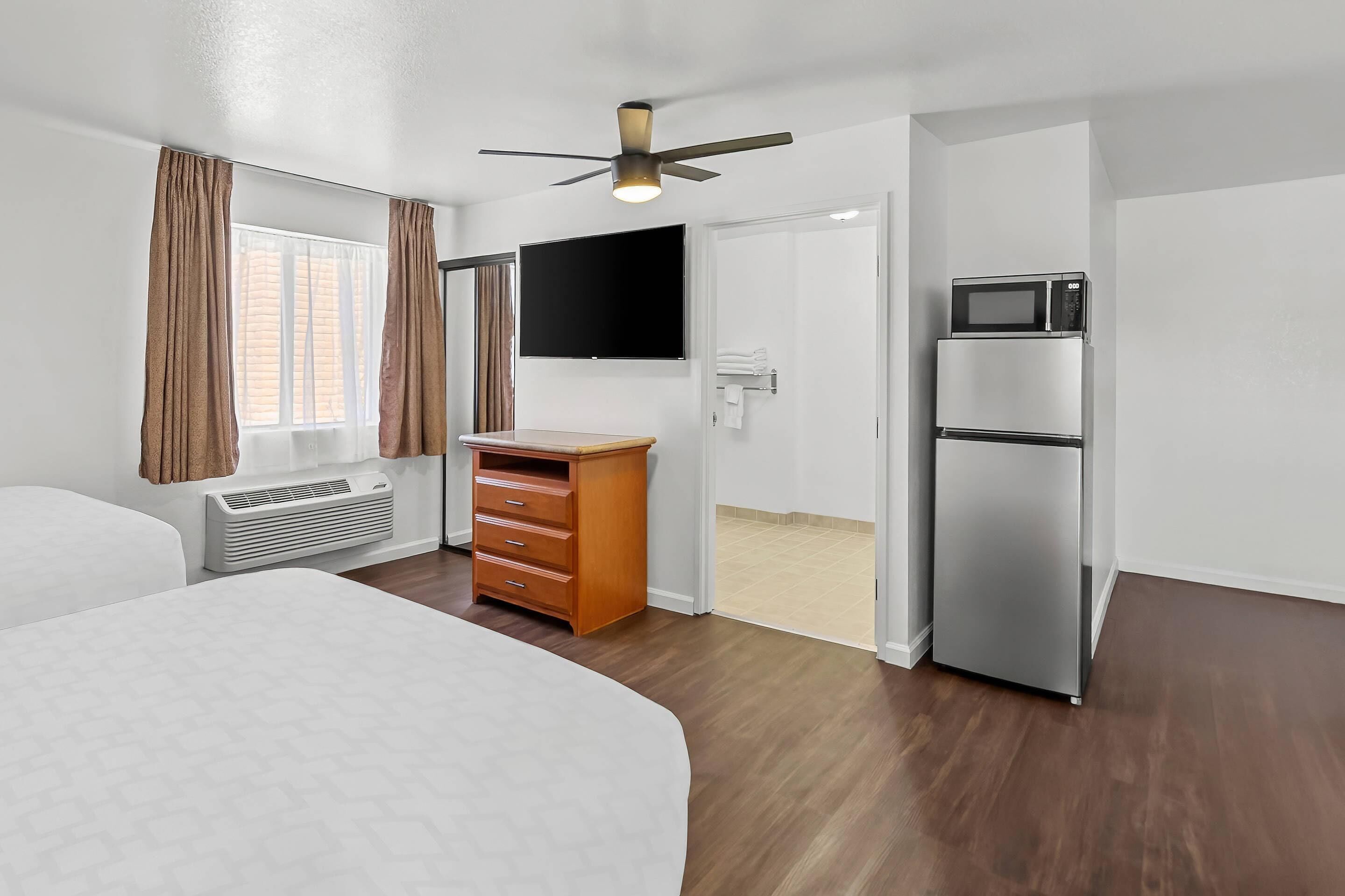 Ein Hotelzimmer mit Bett, Schreibtisch, Kleiderschrank, Flachbildfernseher, Mikrowelle und Deckenventilator.