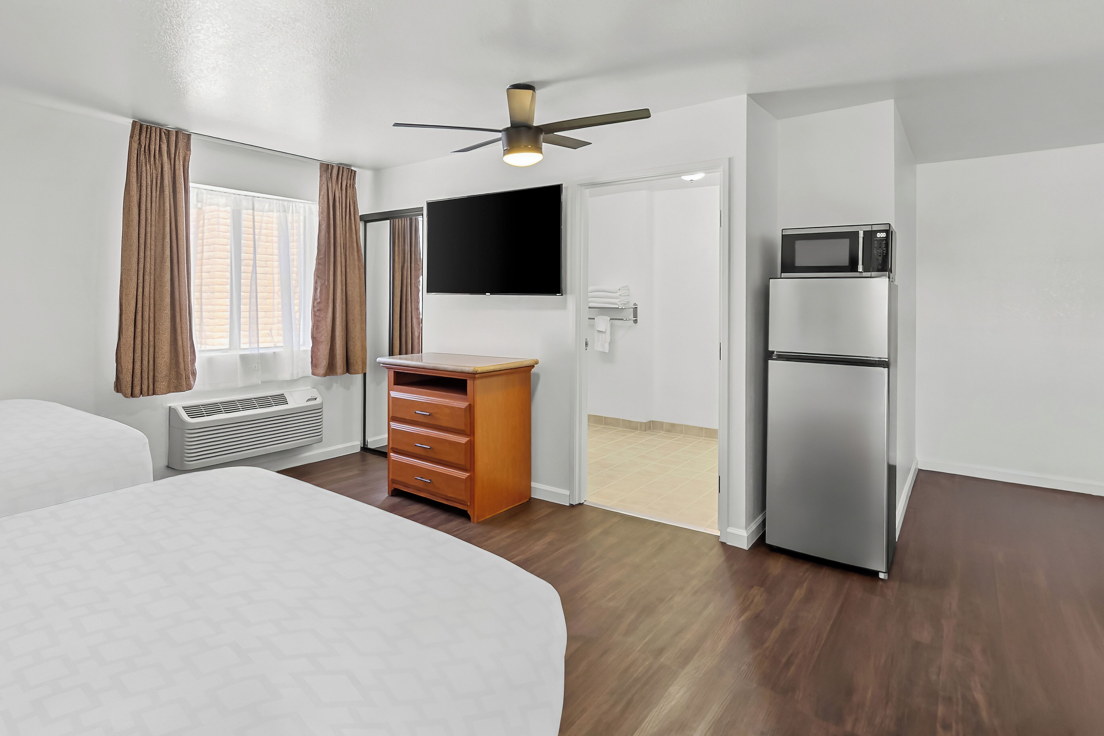 Ein Hotelzimmer mit einem Bett, einem Schminktisch, einer Mikrowelle, einem Kühlschrank, einem Deckenventilator, einem Fernseher und einer Klimaanlage.