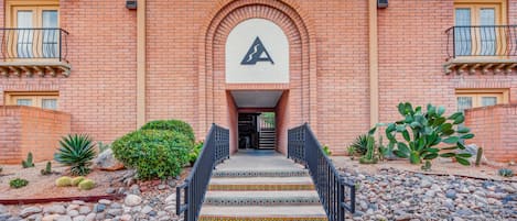 A brick building with a central archway, a staircase leading to a door, and a small garden with cacti and shrubs.