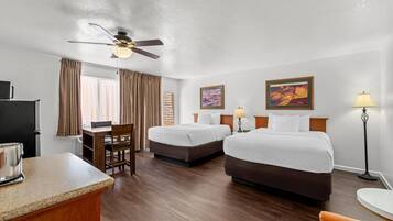 A hotel room with a desk, two beds, a ceiling fan, and artwork on the walls.