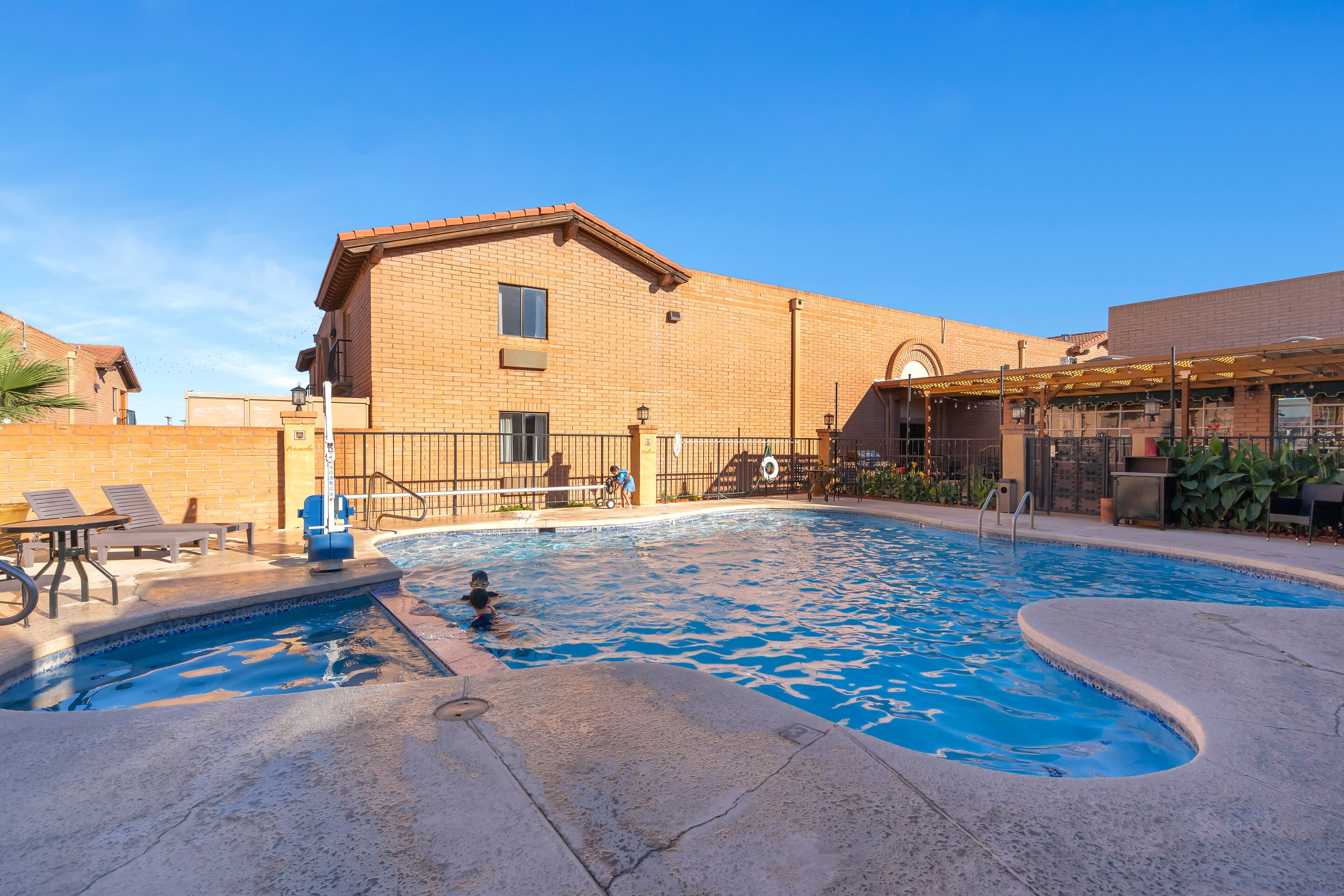A swimming pool area with a brick building, lounge chairs, and a covered patio.