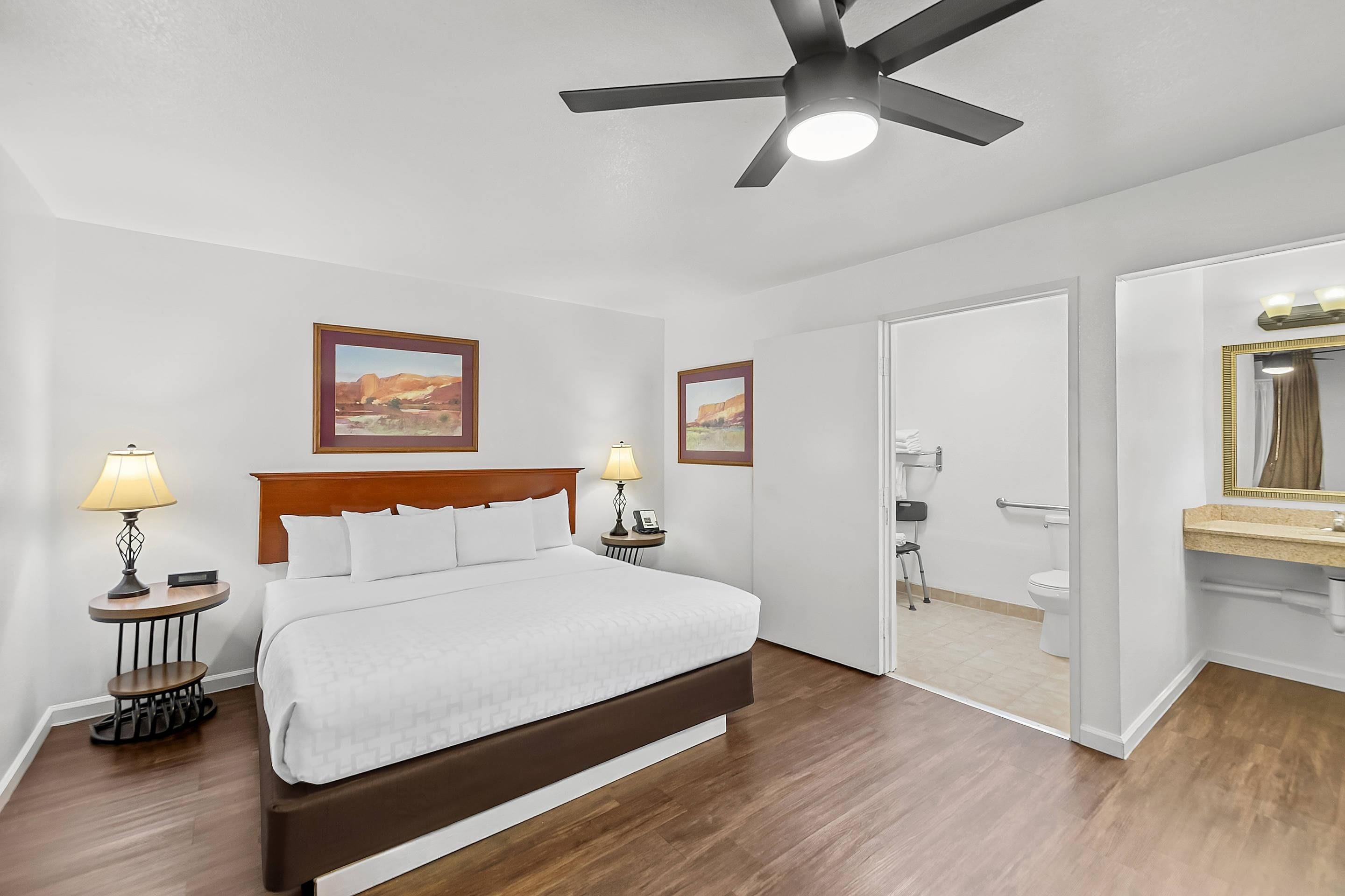 Habitación de hotel con cama, lámparas de noche, ventilador de techo y baño con inodoro y lavamanos.