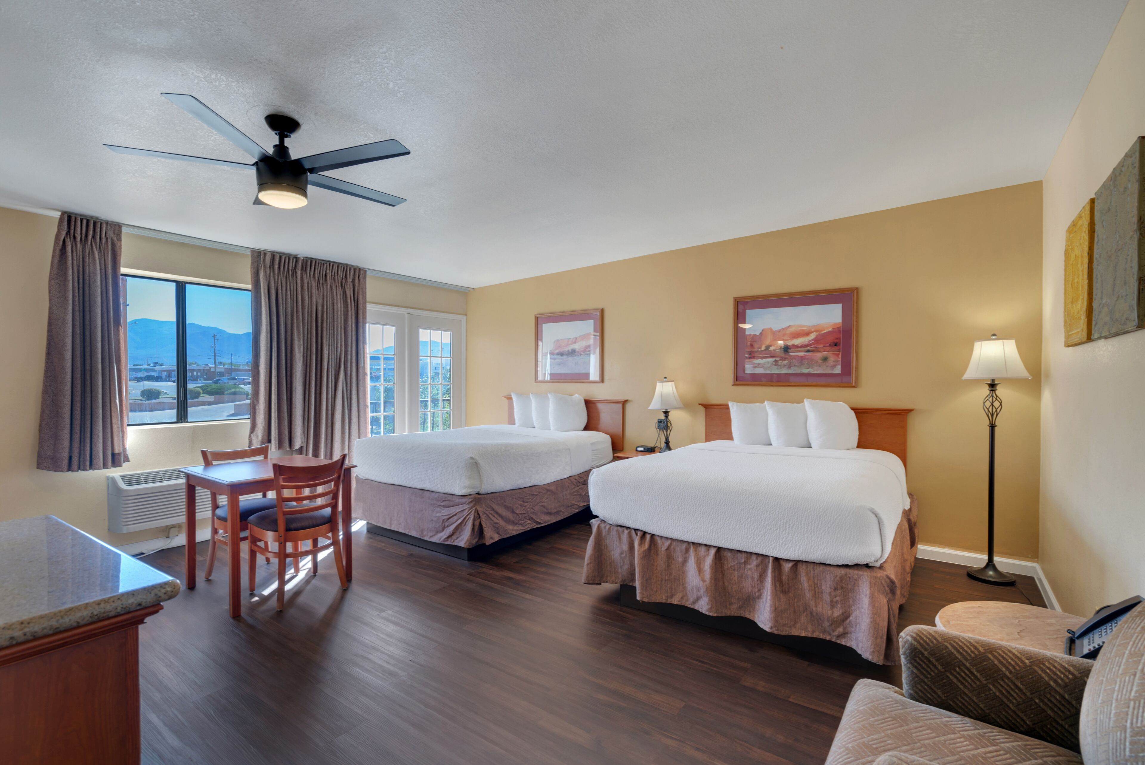 Quarto de hotel com duas camas, uma escrivaninha, uma cadeira, um abajur e vista para as montanhas.