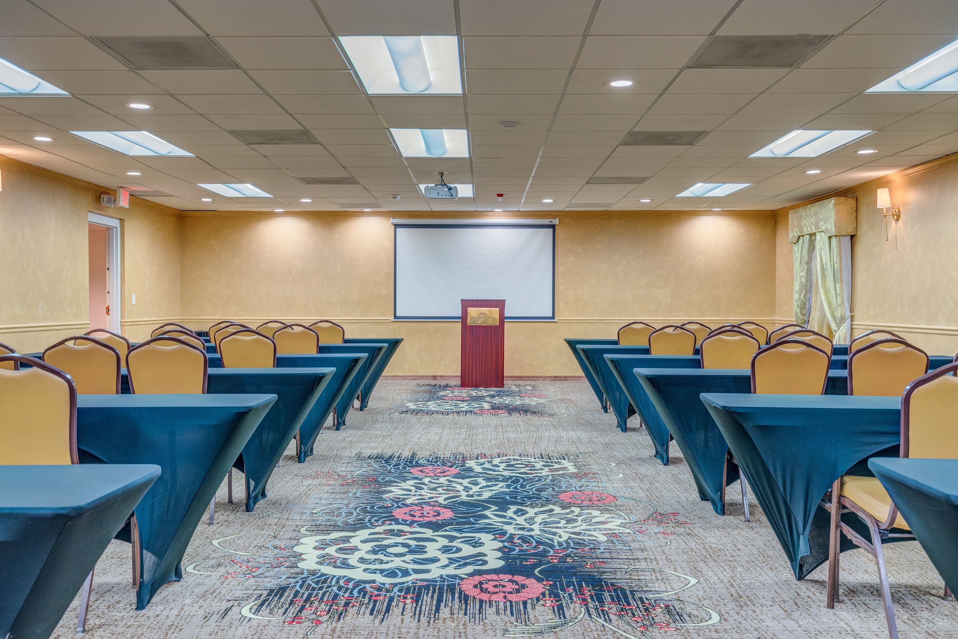 A conference room with rows of tables and chairs, a projector screen, and a carpeted floor.