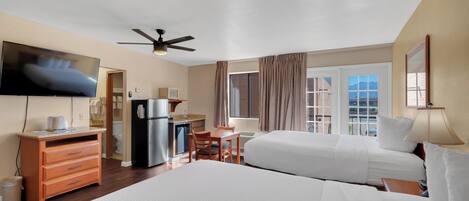 A hotel room with two beds, a ceiling fan, a TV, a kitchenette, and a window with curtains.