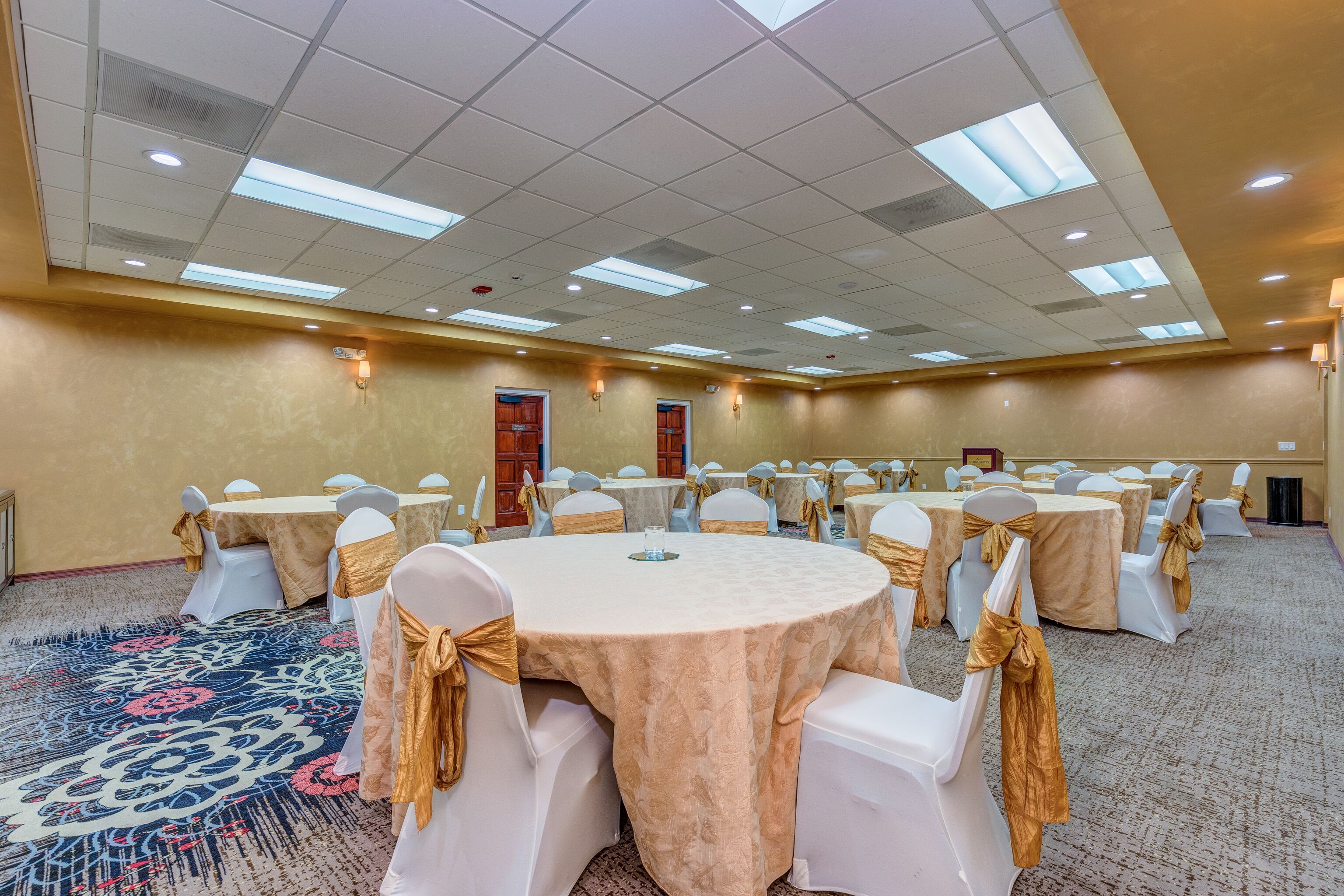Un salón de conferencias con mesas redondas, sillas blancas y fajines dorados.