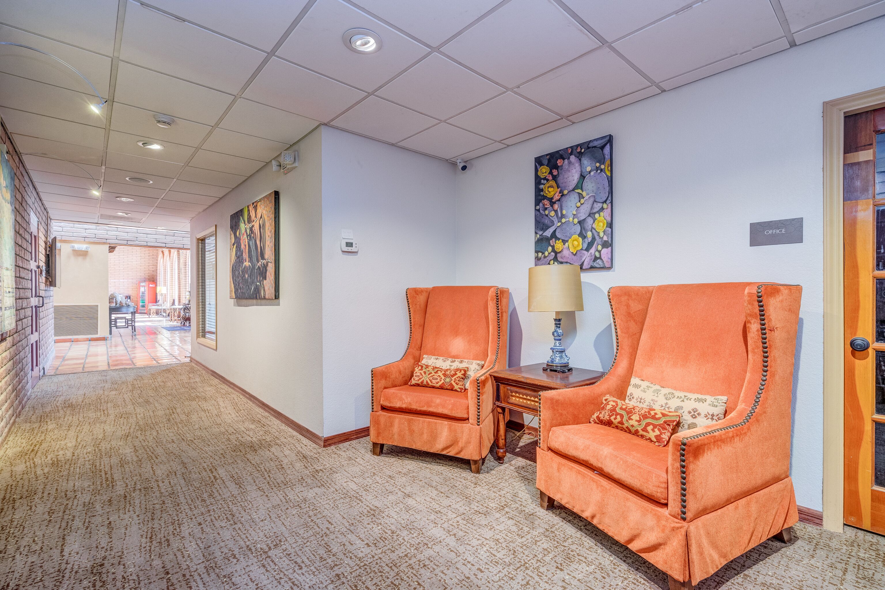 Een hotel lobby met een vloerbedekking, twee oranje fauteuils met decoratieve kussens, een kleine tafel met een lamp en een schilderij aan de muur.