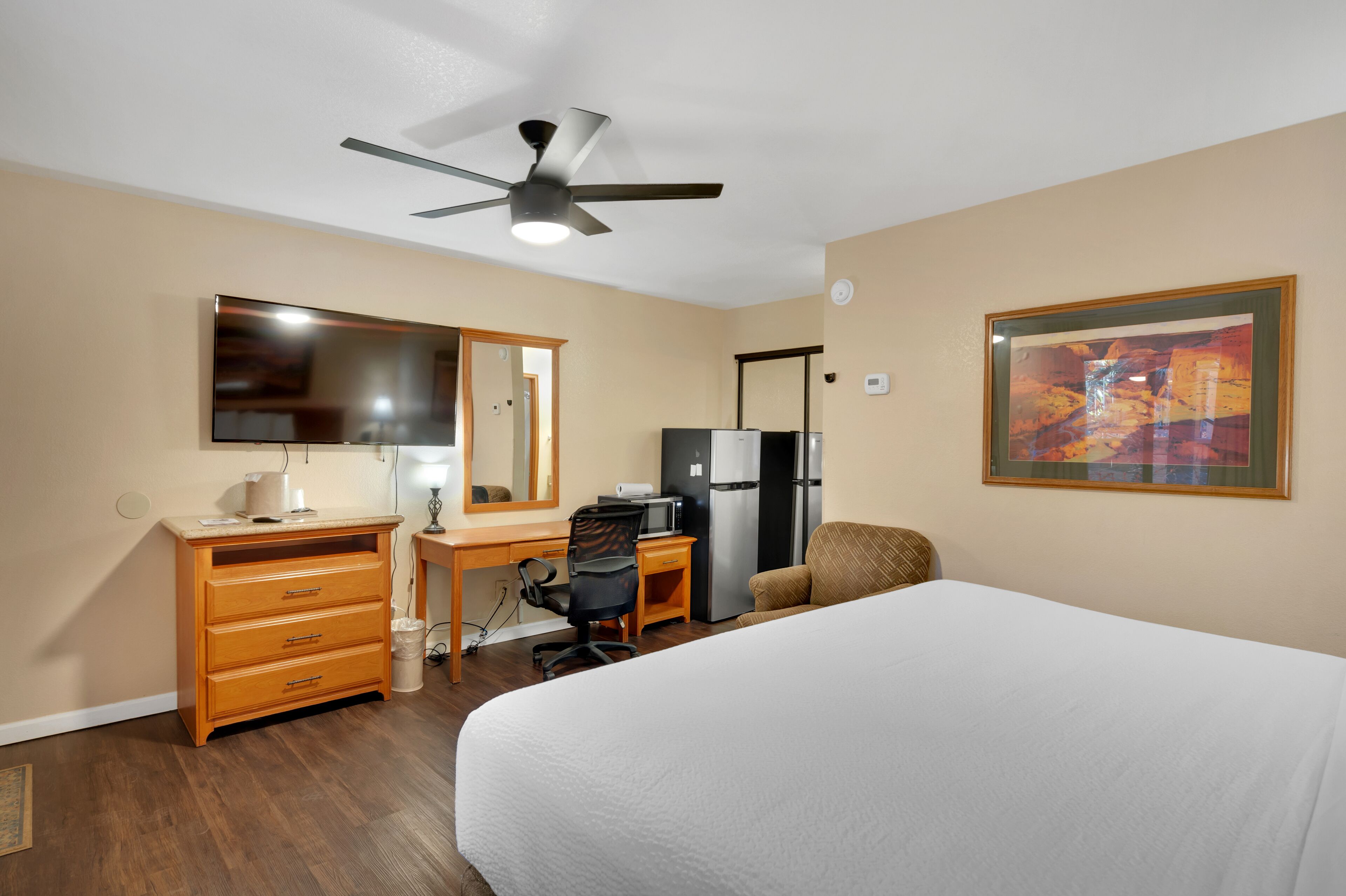 Habitación de hotel con cama, escritorio con silla, refrigerador, microondas, ventilador de techo, espejo grande y un cuadro en la pared.