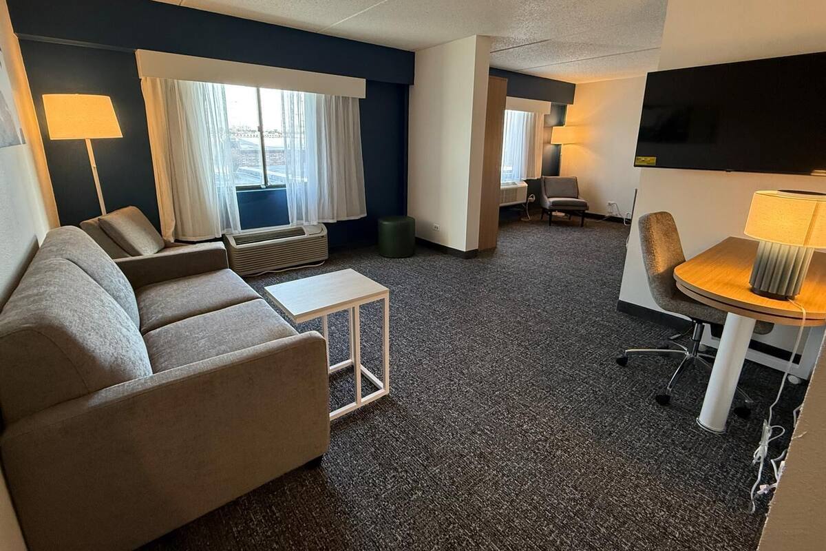 Habitación de hotel con sofá, una mesita, una silla, televisión y ventana con cortinas.