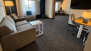 A hotel room with a sofa, a small table, a chair, a TV, and a window with curtains.