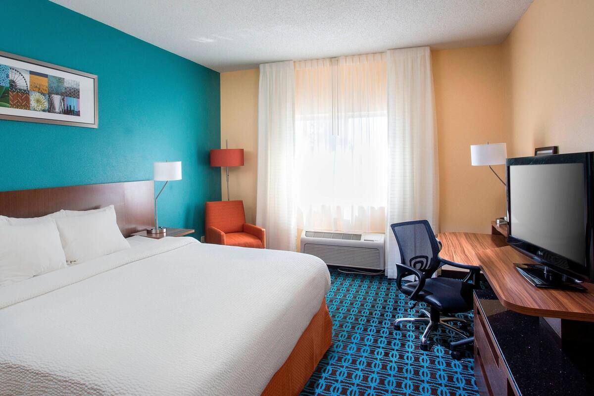 A hotel room with a large bed, a desk with a computer, a TV, and a window with curtains.