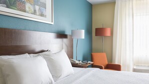A hotel room with a bed, a nightstand, a lamp, an orange chair, and a painting on the wall.