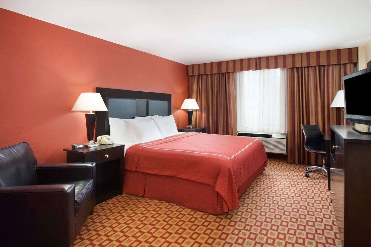 A hotel room with a bed, two bedside tables with lamps, a desk with a television, a chair, and a patterned carpet.