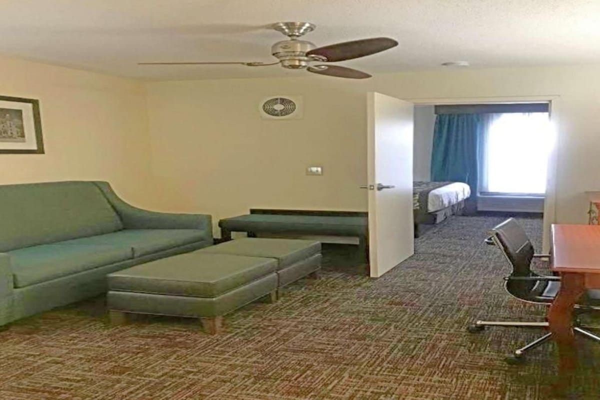 A hotel room with a sofa, ottoman, ceiling fan, and a bed in the adjacent room.