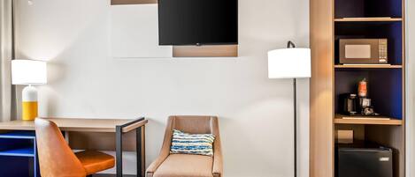 A hotel room with a desk, chair, TV, lamp, and a minibar.