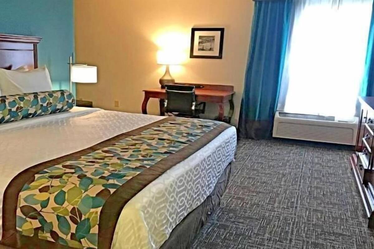 A hotel room with a bed, desk, chair, lamp, and air conditioning unit.