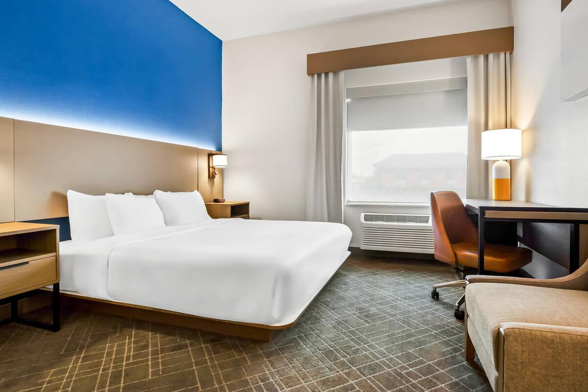 A hotel room with a large bed, a desk with a chair, a window with curtains, and a wall-mounted lamp.