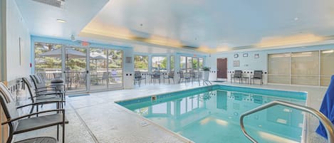 An indoor swimming pool with clear glass walls, a poolside seating area, and a ceiling with recessed lighting.
