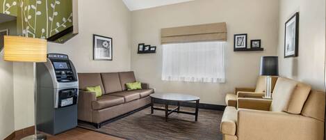 A hotel room with a sofa, armchair, and a small table. There is an ATM machine and a window with curtains.