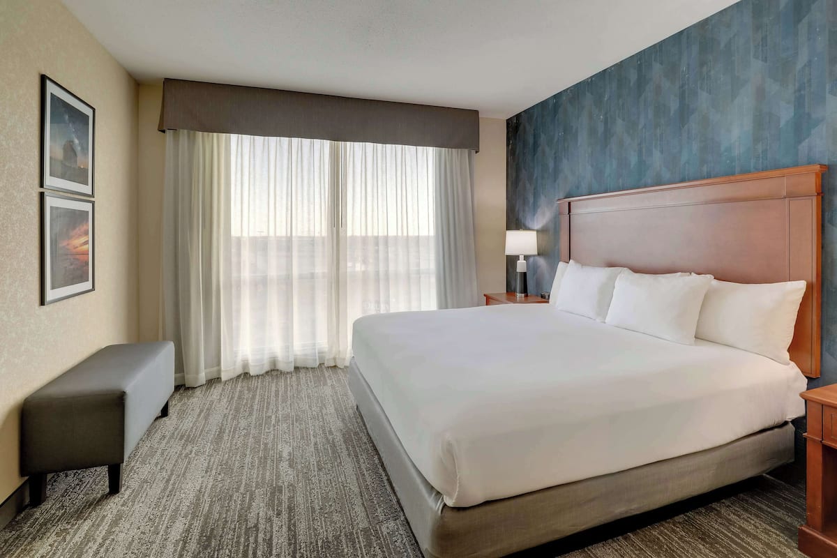 A hotel room with a large bed, a bench, a nightstand with a lamp, and a view of the city through sheer curtains.