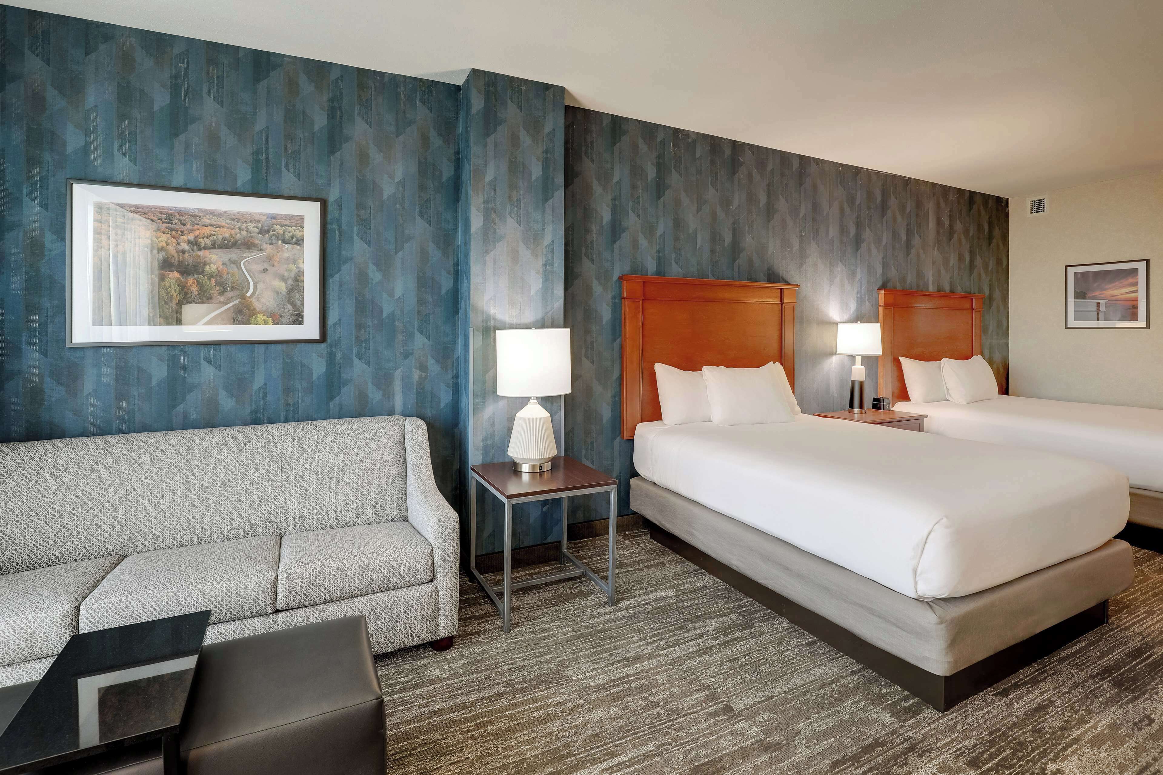 A hotel room with a sofa, two beds, a nightstand, and a lamp.