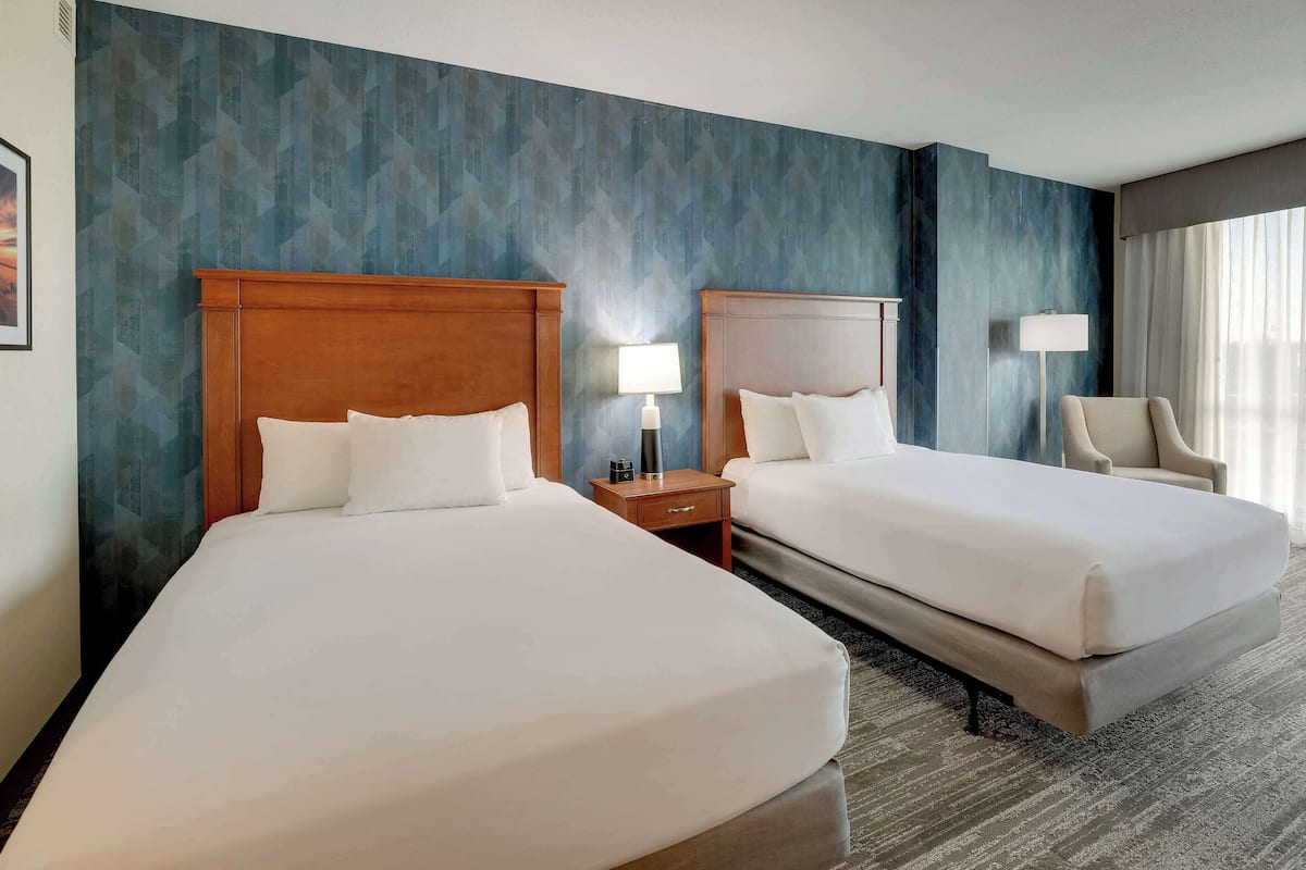 A hotel room with two beds, a nightstand with a lamp, a chair, and a large window with curtains.