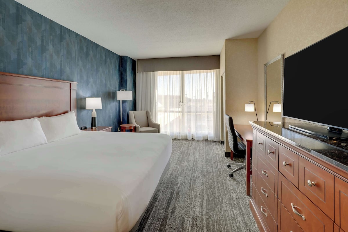 A hotel room with a large bed, a desk with a flat-screen TV, a chair, a desk lamp, and a window with sheer curtains.