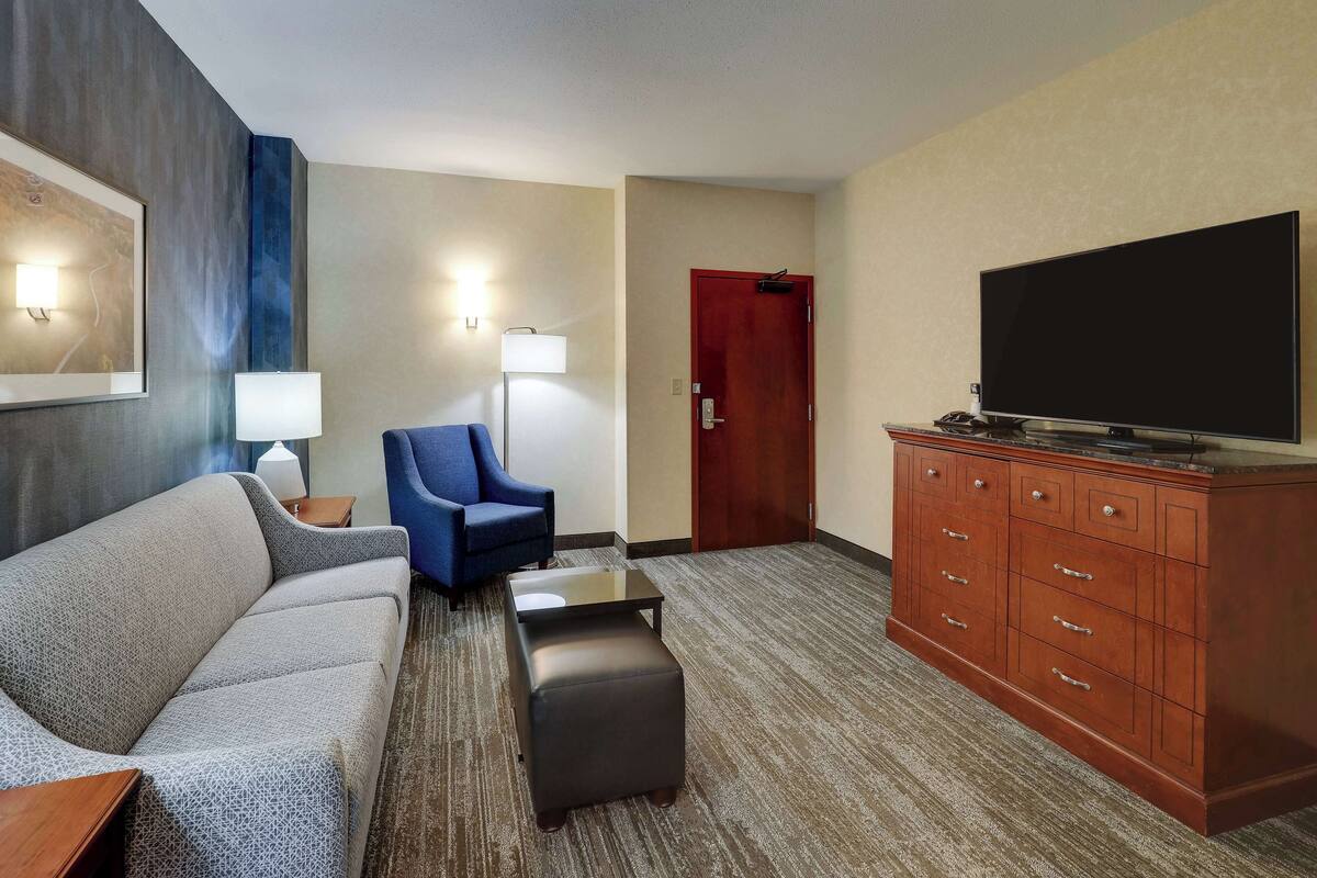 A hotel room with a sofa, ottoman, blue armchair, a flat-screen TV, a wooden cabinet, and a lamp.