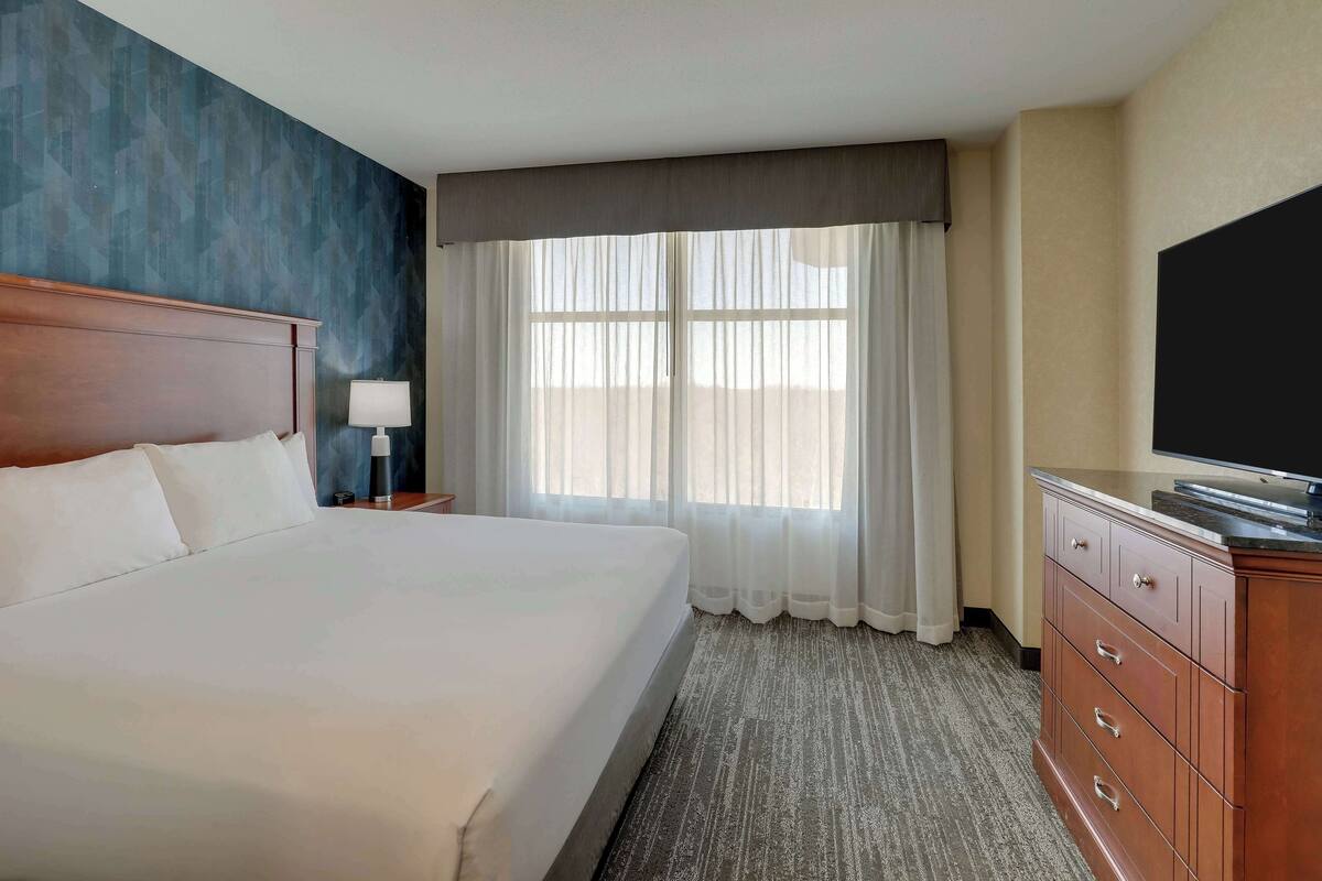 A hotel room with a large bed, a wooden headboard, a dresser, a flat-screen TV, and a window with sheer curtains.