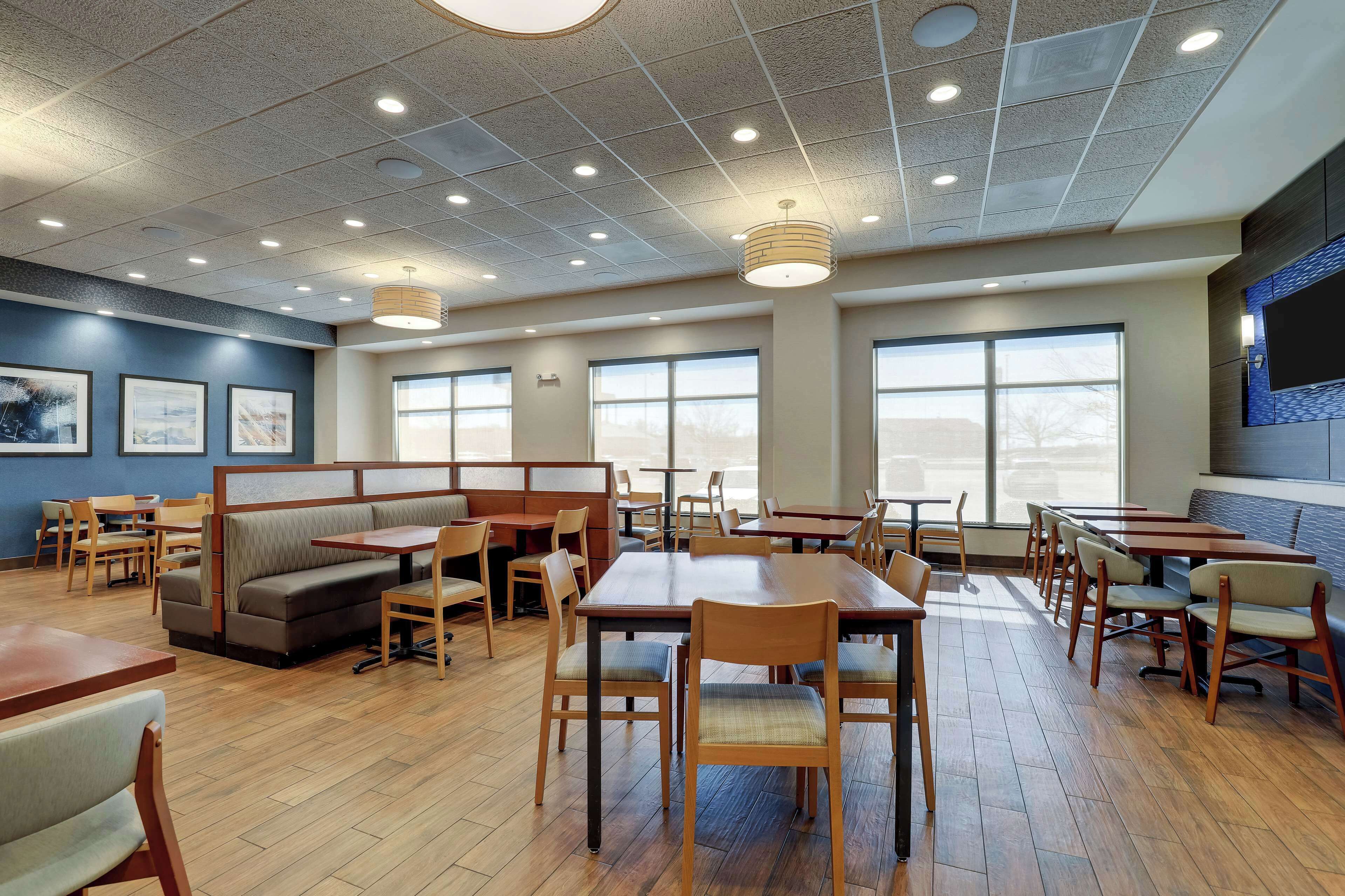 Un comedor amplio con mesas y sillas de madera, un área de estar central con un banco, y ventanas grandes que permiten la entrada de luz natural.