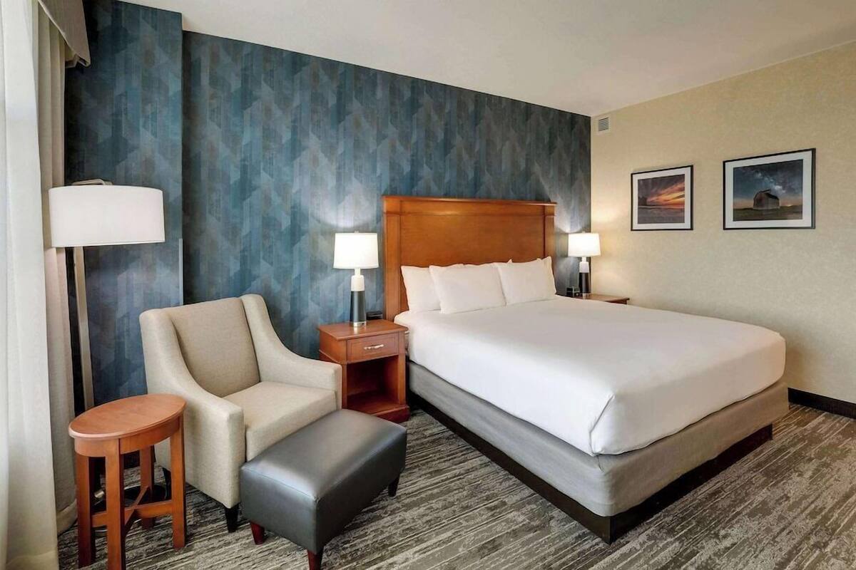 A hotel room with a large bed, two bedside tables with lamps, a chair, a small stool, and two framed pictures on the wall.