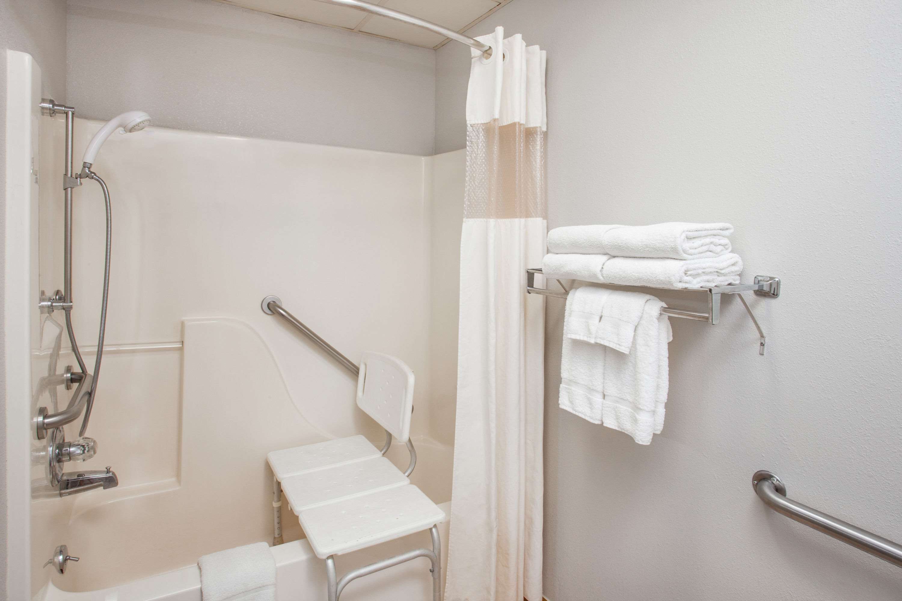 A bathroom with a shower, a grab bar, a towel rack with folded towels, and a white chair.