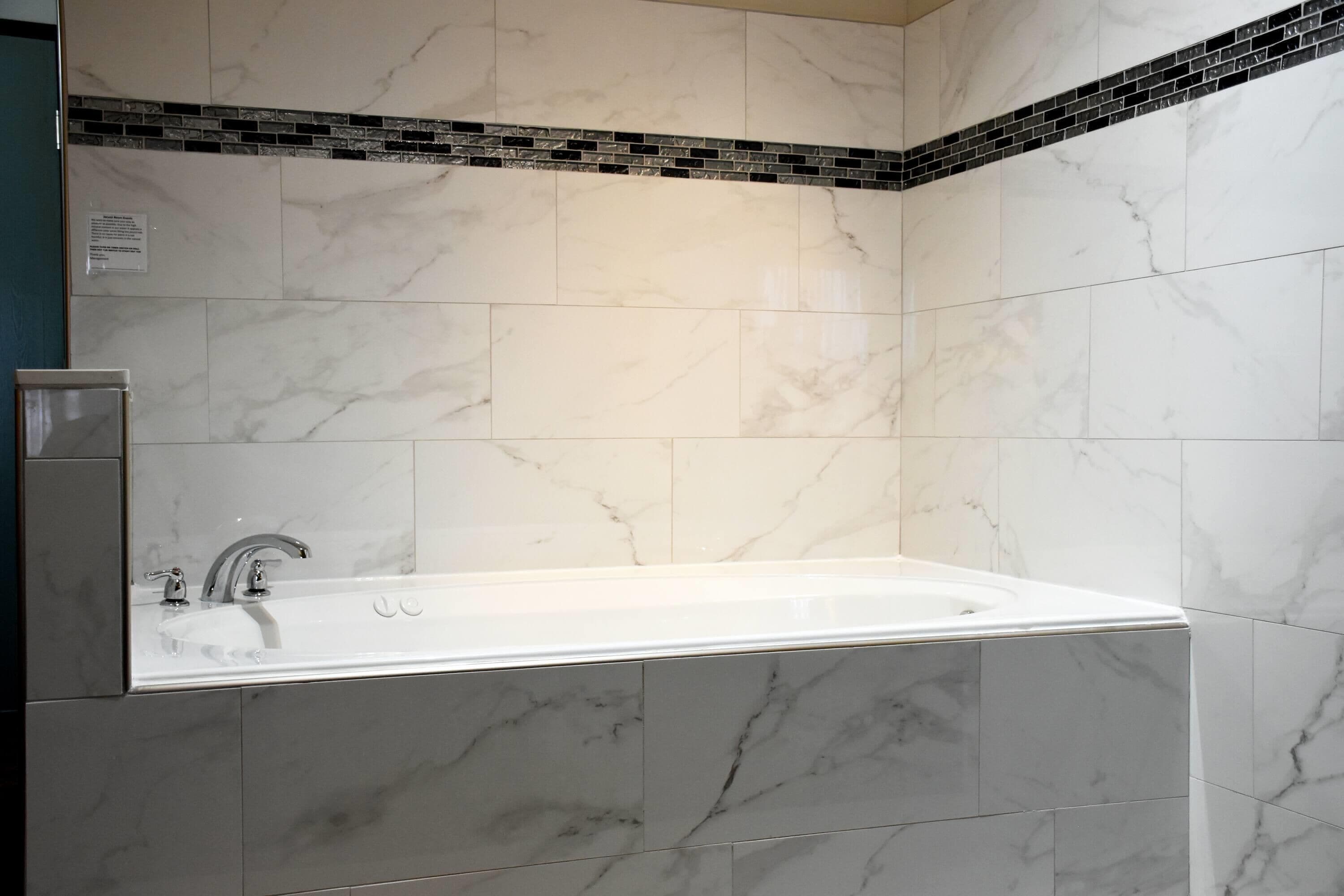 A bathroom with a large white bathtub, grey marble tiles, and a black and white mosaic border.