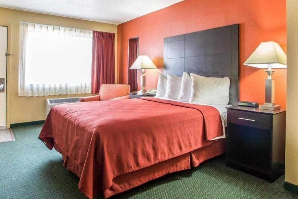 Habitación de hotel con una cama grande, dos mesitas de noche con lámparas, una silla y una ventana con cortinas.