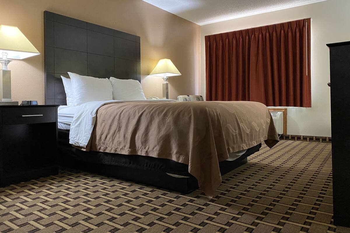 Habitación de hotel con cama, dos mesitas de noche con lámparas, cabecera, ventana grande con cortinas y alfombra estampada.