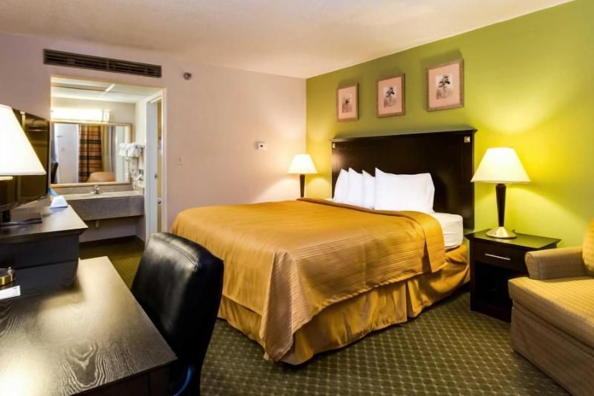 A hotel room with a bed, two bedside tables with lamps, a desk with a chair, a mirror, and a green wall with three framed pictures.