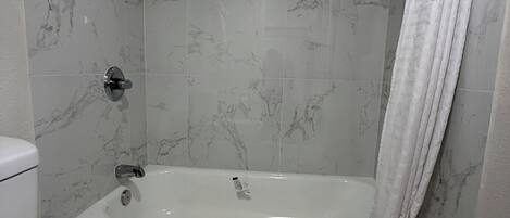 Une salle de bain avec des toilettes blanches, une baignoire équipée d’un pommeau de douche et une serviette accrochée au mur.
