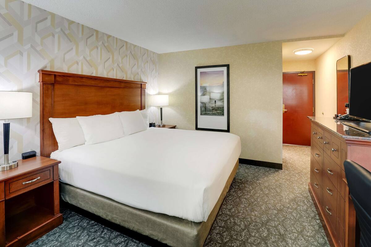 Quarto de hotel com uma cama grande, cabeceira de madeira, mesas de cabeceira com luminárias, penteadeira e um quadro na parede.
