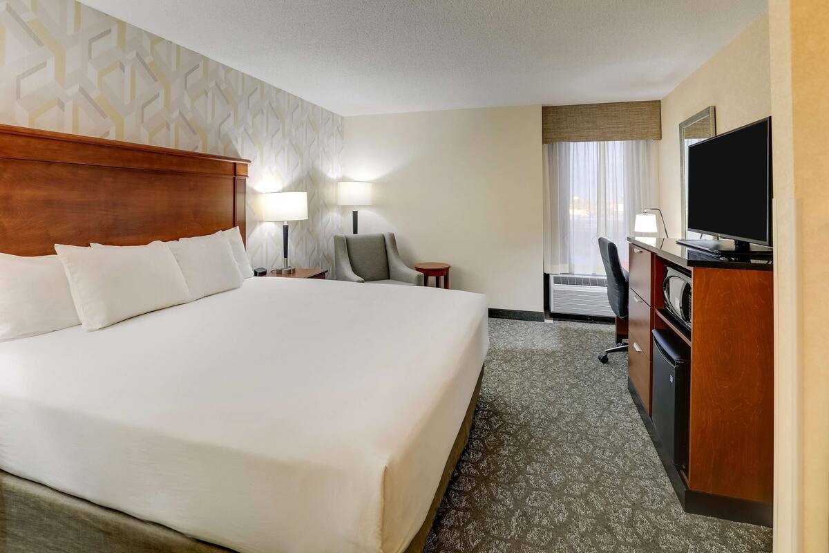 Quarto de hotel com uma cama grande, uma escrivaninha com televisão de tela plana, uma cadeira, um abajur e uma pequena mesa.