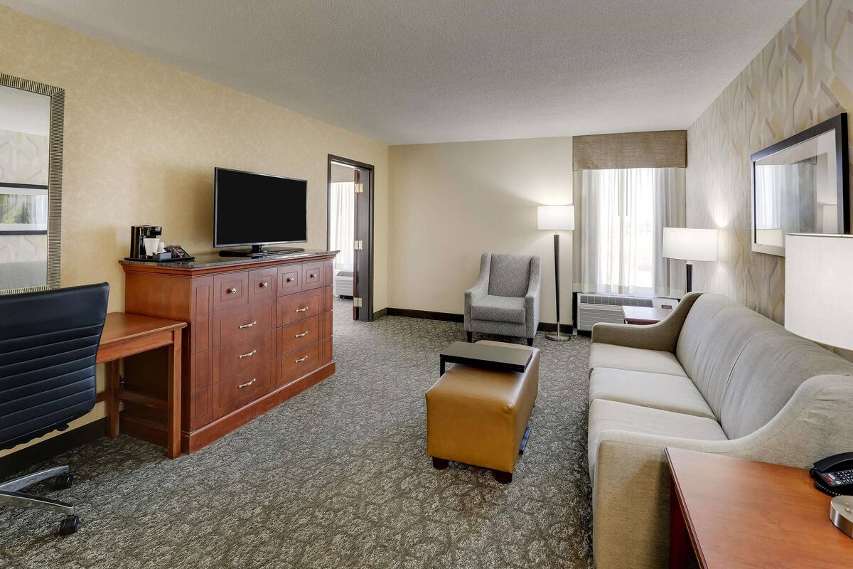 Quarto de hotel com televisão de tela plana, escrivaninha de madeira, poltrona cinza, sofá bege e um pequeno banquinho.