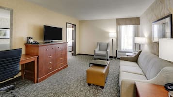A hotel room with a flat-screen TV, a wooden desk, a grey armchair, a beige sofa, and a small ottoman.