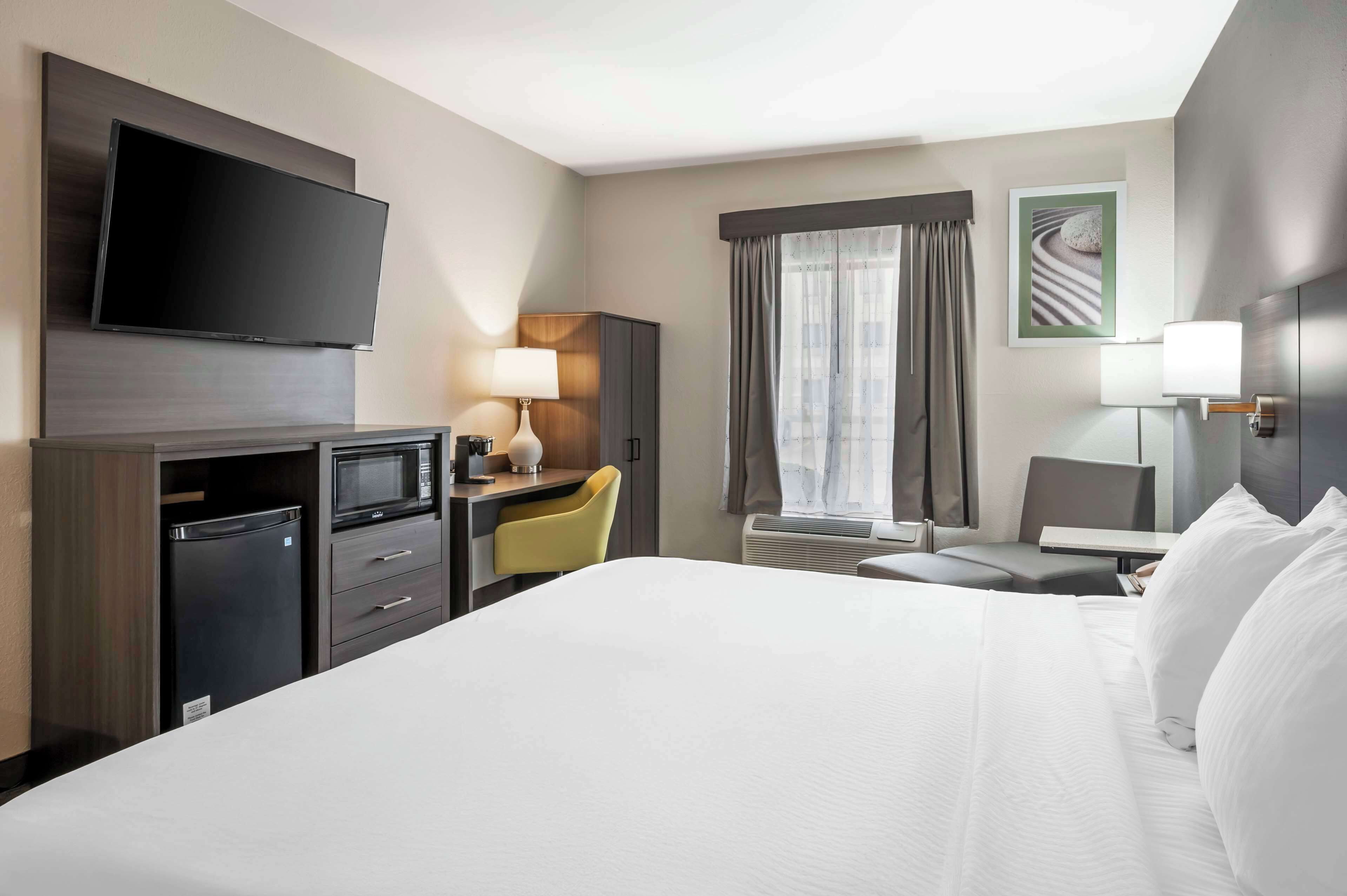 A hotel room with a large bed, a desk with a chair, a flat-screen TV, a microwave, a refrigerator, and a window with curtains.