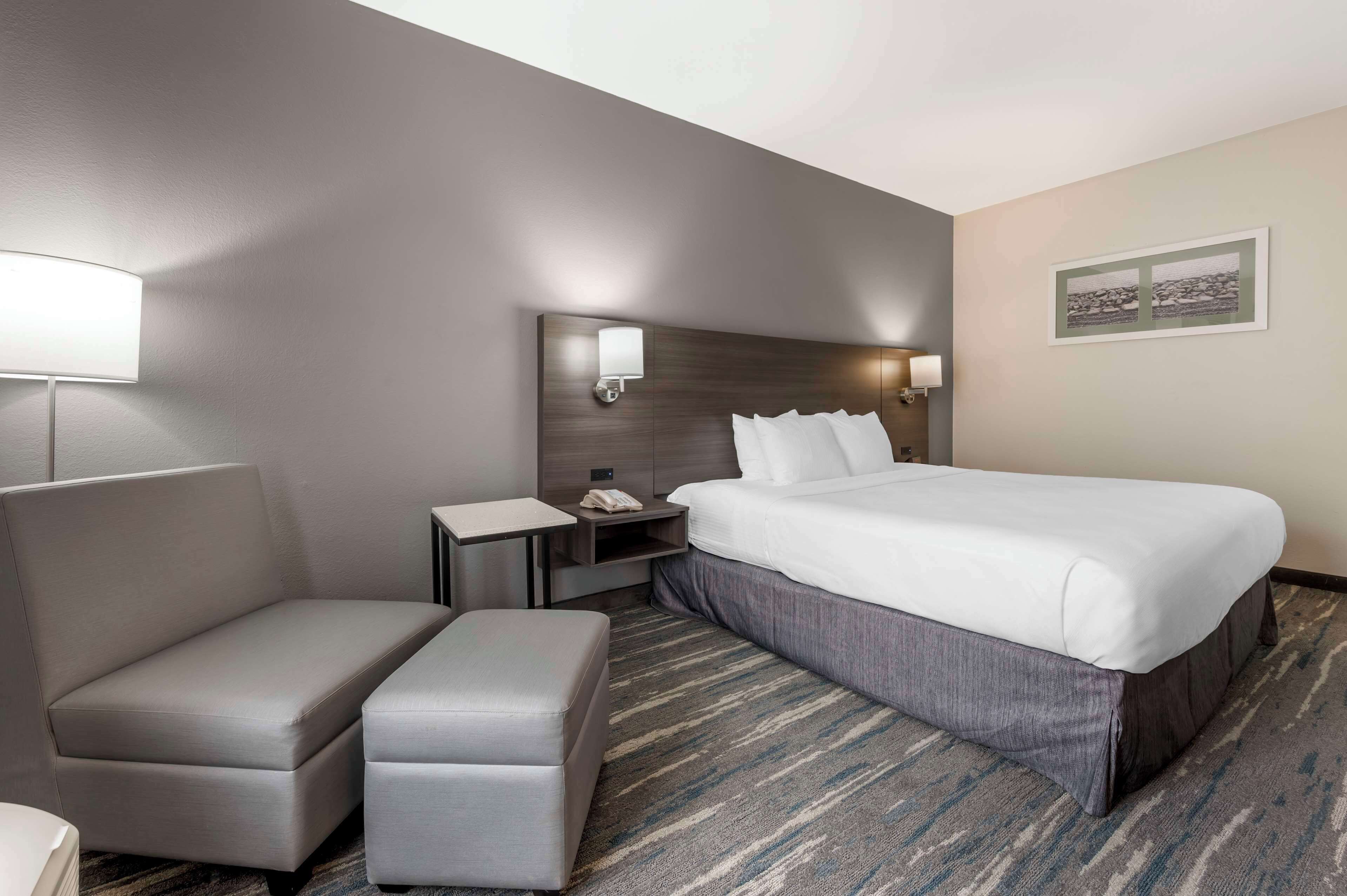 A hotel room with a large bed, a bedside table, a lamp, a chair, and a small ottoman.