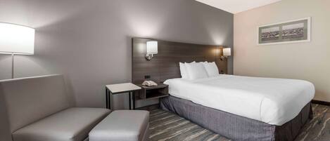 A hotel room with a large bed, a bedside table, a lamp, a chair, and a small ottoman.