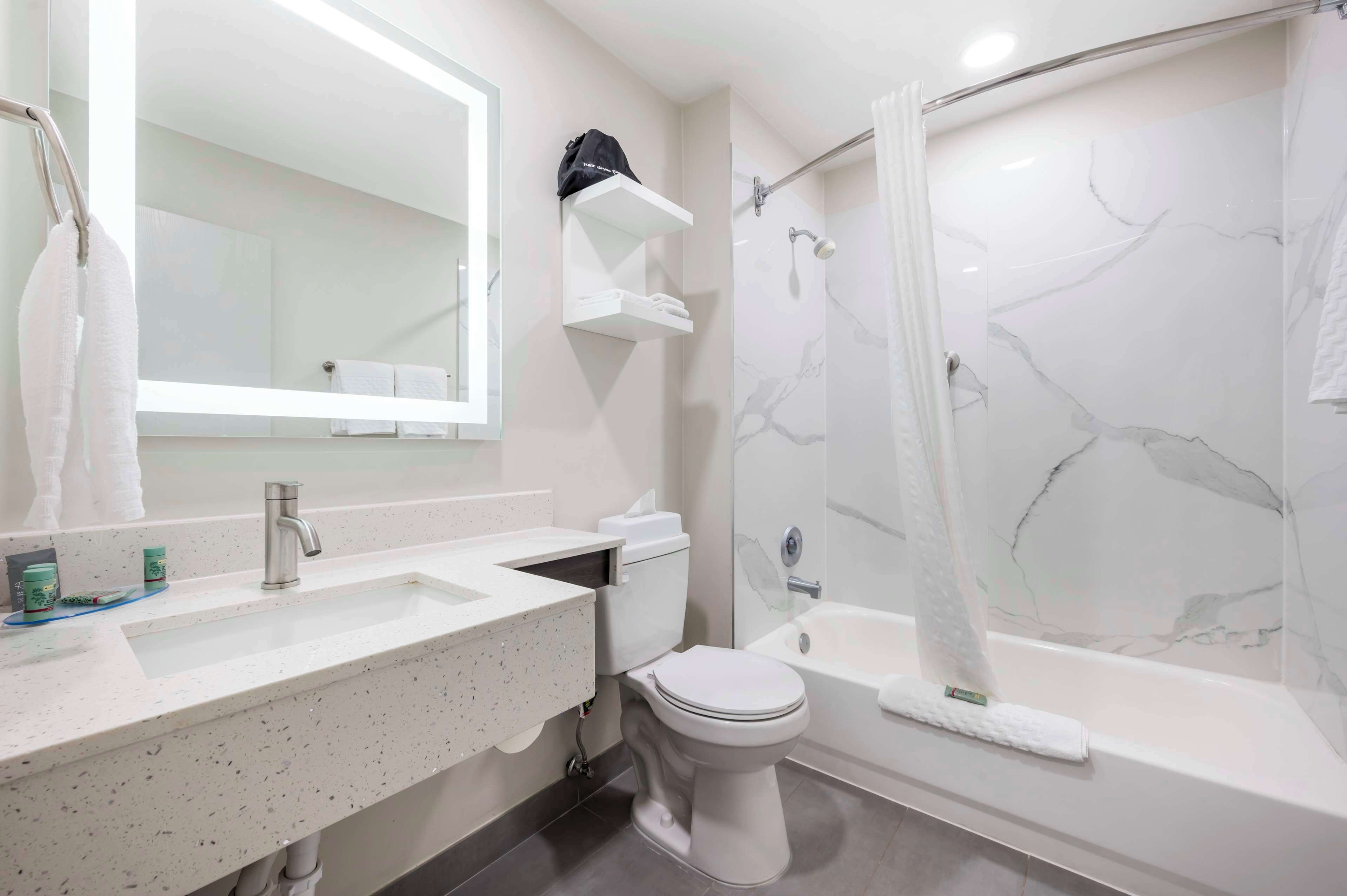 Une salle de bain moderne avec un lavabo, des toilettes et une baignoire, tous de couleur blanche. Un miroir est placé au-dessus du lavabo et un porte-serviettes est fixé au mur.